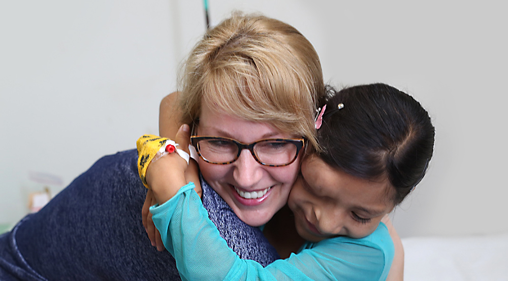 Jessyka Lightall博士在2017年在秘鲁的一名年轻女性患者中收到了一个拥抱。年轻的女孩在她的额头和鼻子上有肿块，在她的手中，胶带覆盖着一个四个港口。她闭着眼睛微笑着。莱德博士微笑着。在背景中，看到了一张医院床。