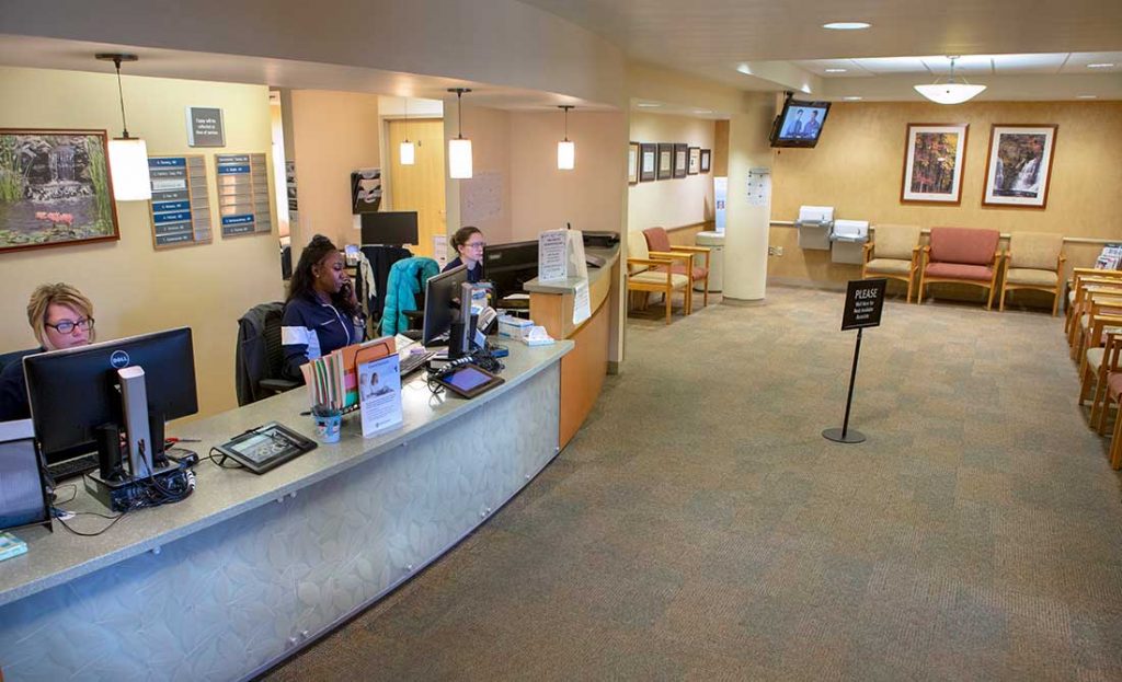 三名妇女工作人员坐在位于位于Penn State Health Milton S. Hershey Medical Centre的面部神经障碍诊所的主桌旁。118金宝搏抽水女性看着他们的电脑显示器。这一部分大堂设有地毯，10把椅子，墙上的艺术品和悬挂式灯具。