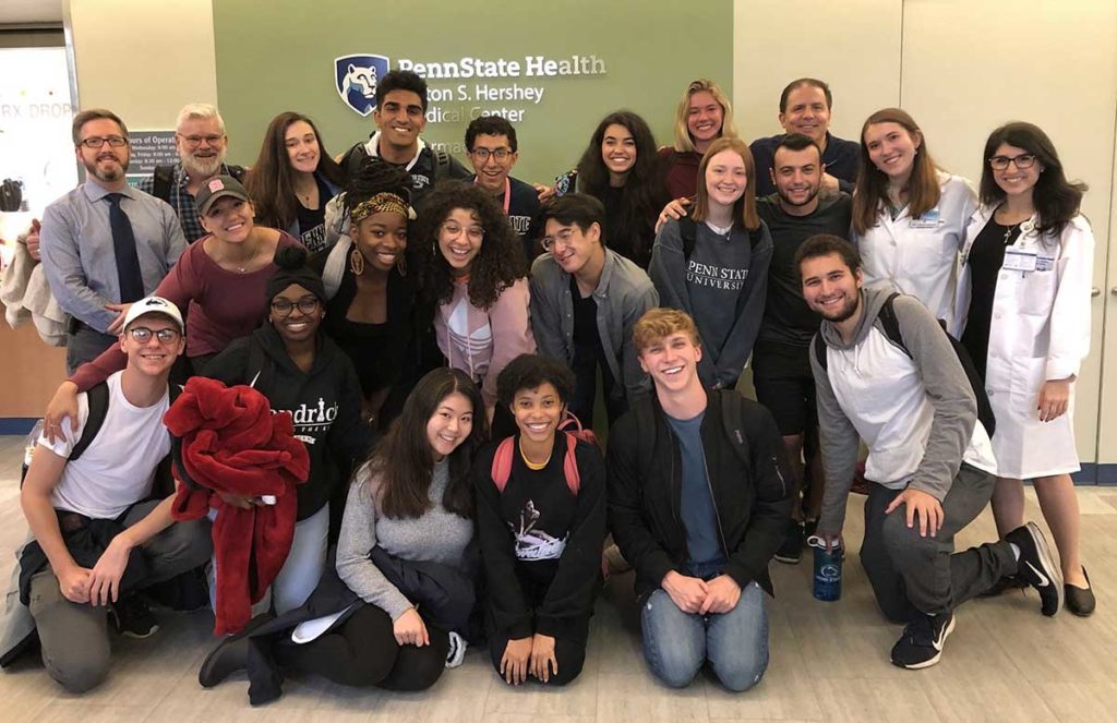 A group of students from the Penn State Department of Musical Theatre stand with Dr. John Gniady, otolaryngologist, in front of a green wall with the Penn State Health Milton S. Hershey Medical Center logo in the background.