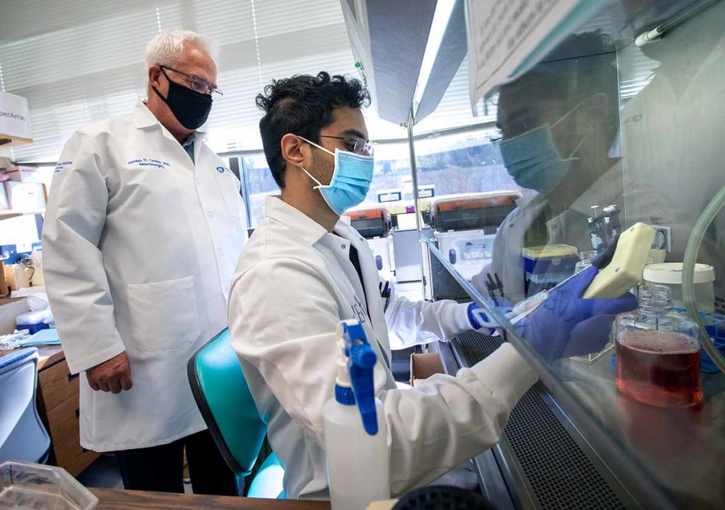 James Connor, distinguished professor of neurosurgery and anatomy and vice chair for research, Department of Neurosurgery, stands in his lab and looks over the shoulder of graduate assistant Ganesh Shenoy. Both are wearing face masks and white lab coats. Shenoy is holding a yellow device and wearing purple gloves.