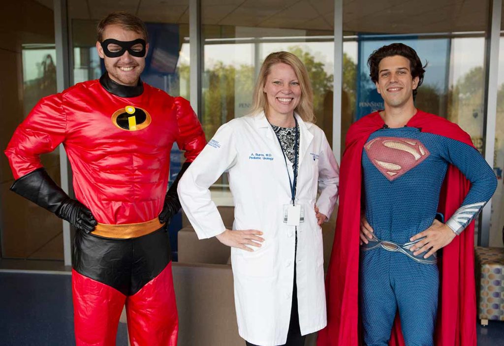 Medical students dressed as Mr. Incredible and Superman flank blonde physician wearing a white lab coat.