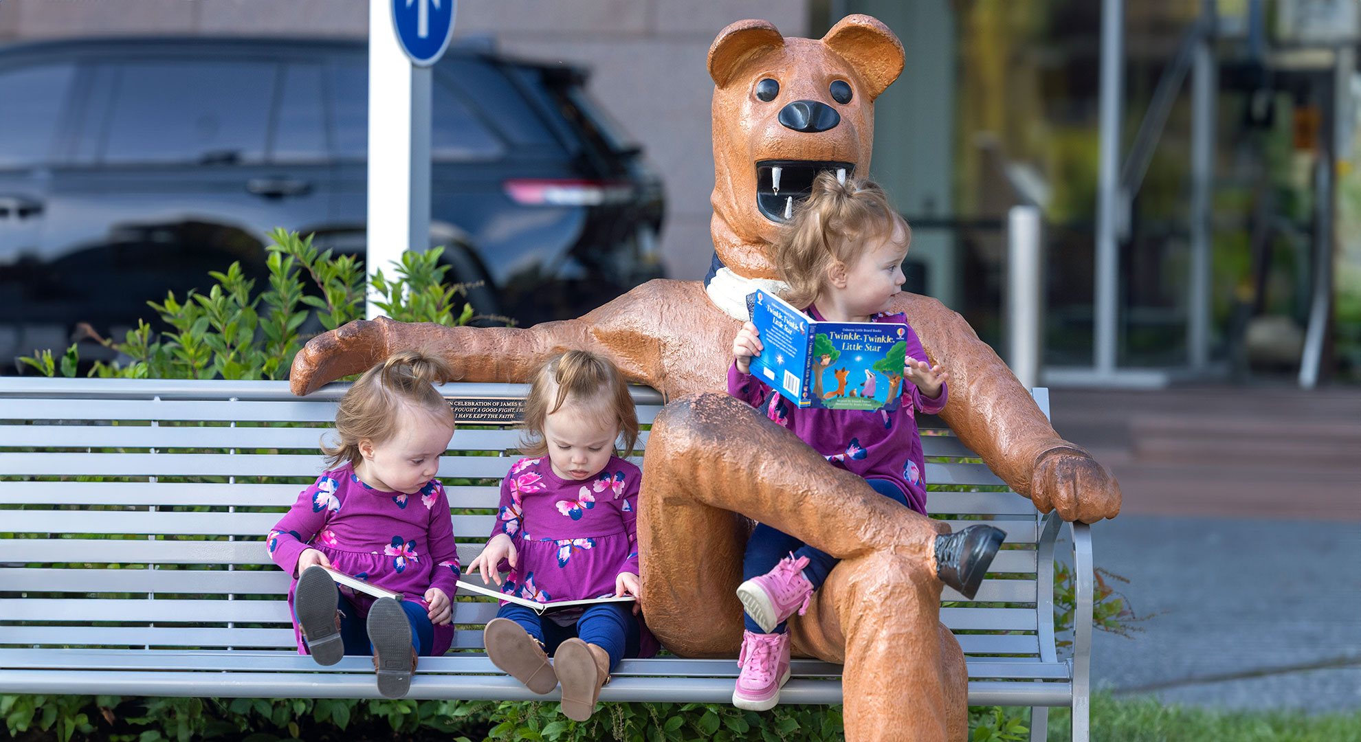 A set of triplet girls, including one seated on the lap of the Nittany lion statue, sits on a bench reading books. On the left behind the bench, the top of a plant, a pole and a car are visible. To the right, is the building entrance.