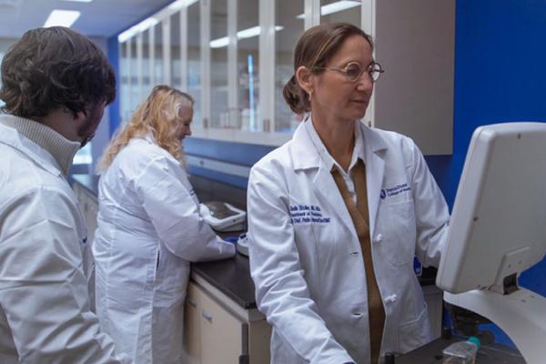 Dr. Giselle Saulnier-Sholler looks at a screen while working on research in her lab while two members of the lab also work.