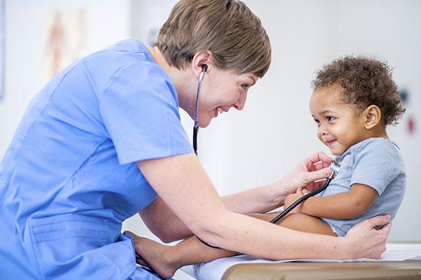 Side view of a young mixed race baby sitting up on an exam table in a gray  diaper shirt. He has a smile on his face as the female doctor holds his waist and listens to his heart with her stethoscope. The doctor is dressed in blue medical scrubs and is smiling back at the baby while she listens.