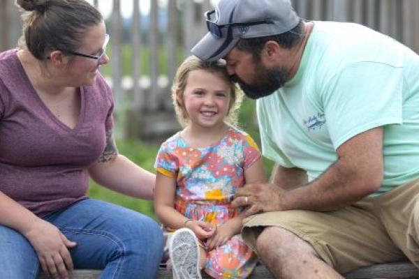 A man and a woman to the left and right of a little girl in a dress on a park bench. The little girl smiles as the man leans down and nuzzles the side of her head.