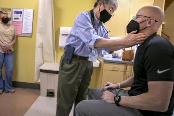 Dr. Shin Mineishi, left, of Penn State Cancer Institute, touches the neck of a patient, Doug Harris in a patient exam room. On the left, Harris’ wife Lisa stands and watches. All three are wearing face masks.