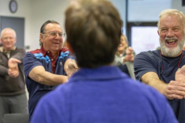 Hank Shipman smiles broadly as he stretches his arms out in front of himself and bends down his fingers. He wears a T-shirt and has a beard and moustache. Other rehab patients smile and laugh in the background as they do similar stretches. The instructor is seen from behind.