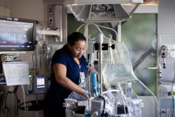 A neonatal intensive care unit (NICU) nurse adjusts equipment and cares for an infant inside an incubator. Medical monitors, IV pumps, and respiratory equipment surround the bedside, while a vital signs monitor displays the baby's heart rate and other readings.