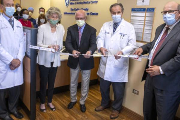 Five people in the foreground pose for a photo as they prepare to cut a ceremonial ribbon. A small group of people looks on in the background.