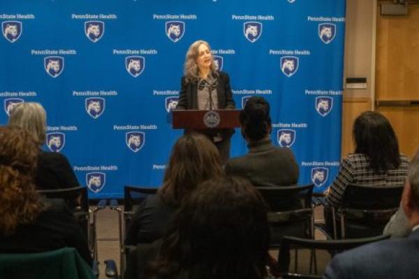 A woman in professional attire speaks at a lectern, gesturing with her hands. A step-and-repeat ‘Penn State Health’ backdrop is in the background.