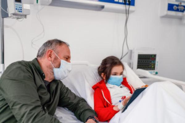 A young girl sits up in her hospital bed, playing on a tablet computer. She’s hooked up to medical equipment. A man seated at the side of the bed looks at the screen with her.