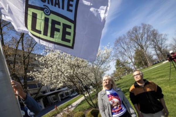 A man and a woman stand in a grassy area looking up at a ‘Donate Life’ flag hoisted several feet into the air on a flagpole. In the background are a sidewalk, a large building, some trees and bushes, and a TV videographer.
