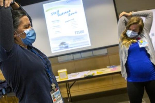 Two women wearing surgical masks and lanyards with nametags pull their elbows as they stretch their arms. A slide with the words “Staff Retreat Zone” is on a screen between them. A table is in the background.