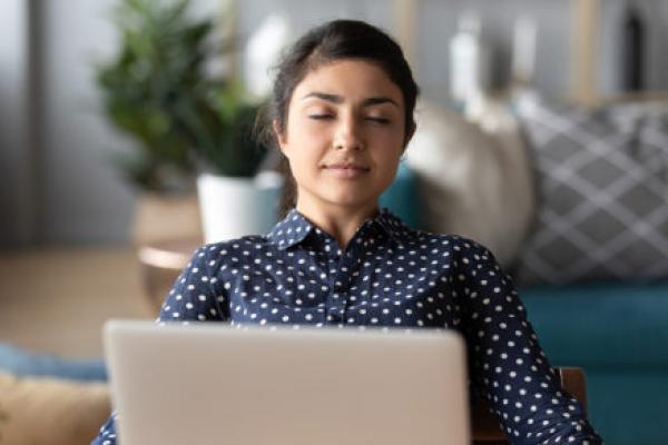 A young woman sits in front of a laptop while meditating in her living room.