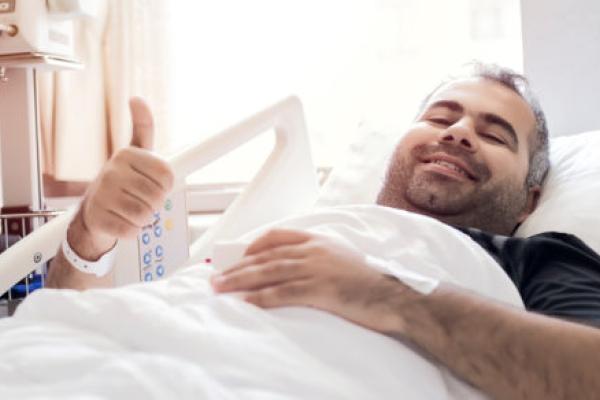 A man lies in a bed, looking up at the camera and giving a thumbs-up with his right hand.