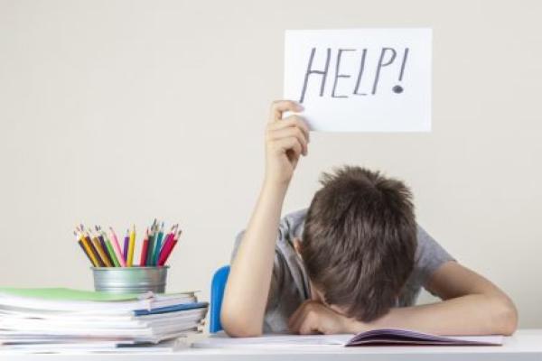 Sad tired frustrated boy sitting at the table with many books and holding help sign.