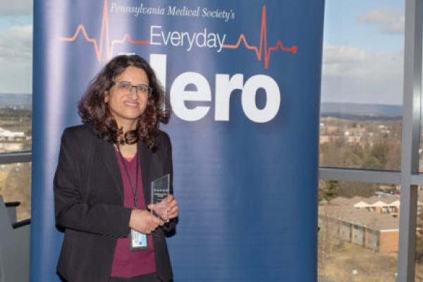 Dr. Gayatra Mainali, wearing professional attire, stands in front of a large pull-up banner with the words “Pennsylvania Medical Society’s Everyday Hero.” Buildings, trees and mountains are in the background, outside a window.