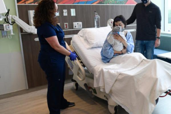 A woman sits in a hospital bed holding a young infant. A medical professional and a man stand at her bedside.