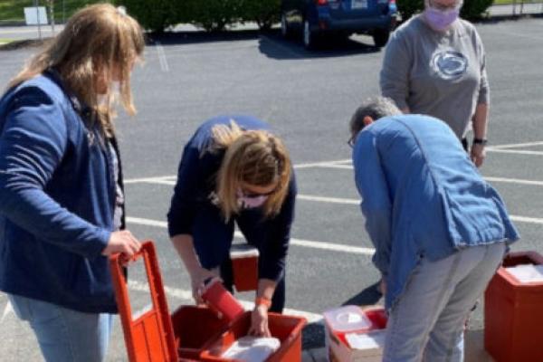 Four health care workers organize plastic containers full of syringes, which are placed at the edge of a parking lot. A car and bushes are in the background.