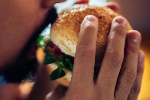 A close-up of a man with a short beard and mustache holding a burger on a bun, about to take a bite out of it. He holds the burger with both hands.