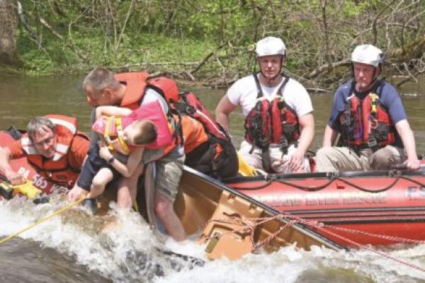 Rex Carmichael, wearing glasses and a life vest, saws tree roots to free a child in a creek as another paramedic holds onto the boy, who is slumped over his arm. Three other paramedics, wearing helmets and life jackets, sit in a river rescue boat.