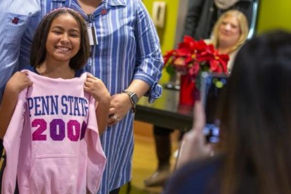 Ruth Vega smiles as she holds a sweatshirt up in front of herself that says Penn State 2000th. Her parents stand behind her. A poinsettia plant sits on a table in the background, with people sitting around it.