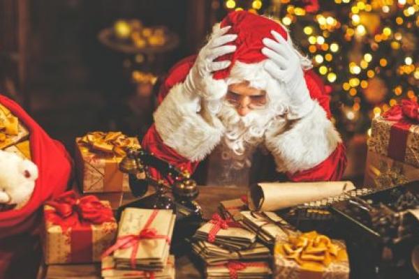 ‘Santa Claus’ rests his elbows on a desk and his head on his hands. The desk is covered with presents and letters. A Christmas tree is in the background, out of focus.