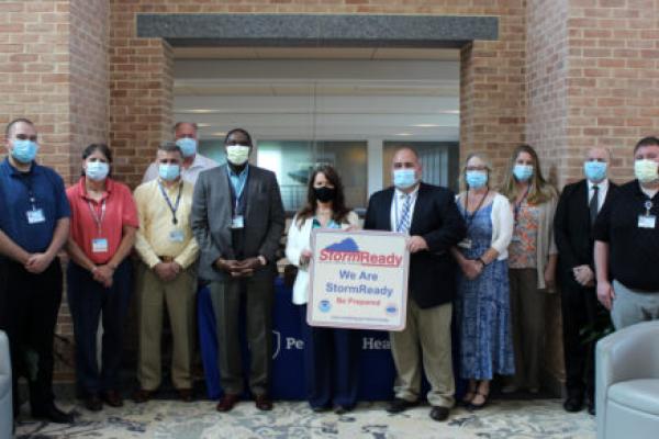 Eleven people stand in a row, posing with a certificate that reads “StormReady: We Are StormReady. Be Prepared.” A brick wall with an opening in the middle is in the immediate background.