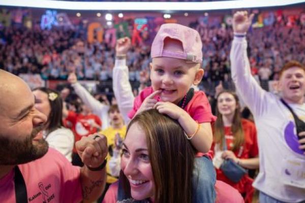 A 2-year-old girl sits on her mom’s shoulders while dad stands to the side, making a smiling face at her. In the background are several hundred people in an arena setting.