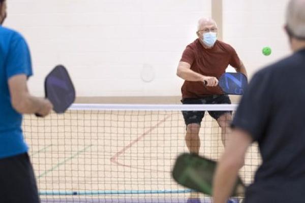 A man in a surgical mask charges a net brandishing a pickleball racquet in a gym. Two other men are seen from behind in the foreground.