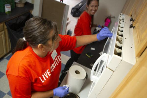 Two women wearing rubber gloves work side-by-side to clean a kitchen in a residential apartment. One looks to the other, smiling.