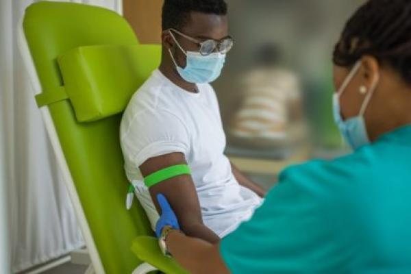 A man sits in a chair at a medical clinic, with a tourniquet on his right arm. A nurse, facing the patient, holds the arm.