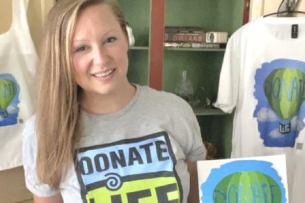 Lauren Gydosh holds the picture she painted of a hot air balloon that says “Donate Life,” surrounded by sky and clouds that contain the words “hope” and “love.” A T-shirt and tote bag bearing the same design hang on cabinet doors behind her. Lauren, who has long, straight hair, is wearing shorts and a T-shirt that says “Donate Life Living Donor.”