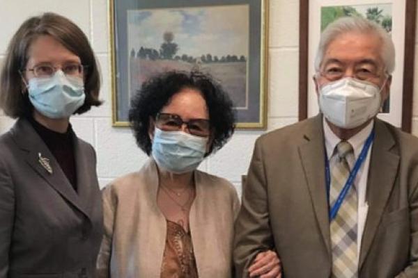 Three people stand next to one another in a room, all wearing face masks. One woman wears a brown suit, and another woman wears a brown jacket. She has her arm linked with a man who is wearing a brown suit and blue checkered tie.