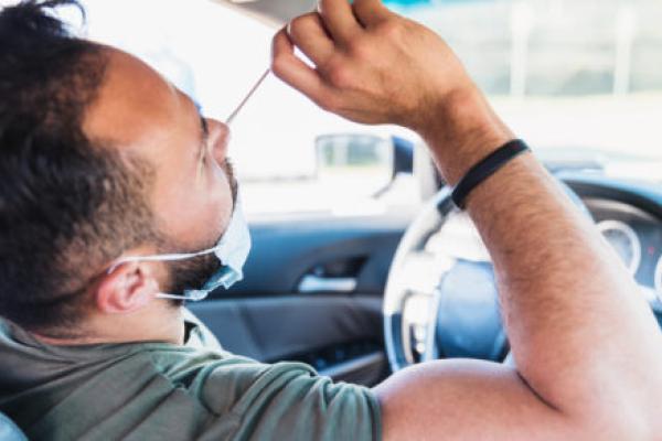 A man sitting in the driver’s seat of a car uses his right hand to insert a nasal swab into his nose, with his head tilted backward slightly.