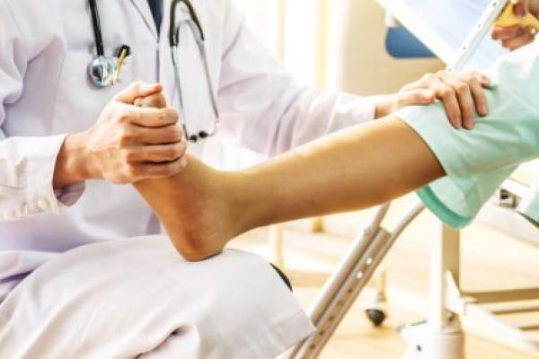 A physician, wearing a medical coat and stethoscope, is seated and holding onto a patient’s lower leg.