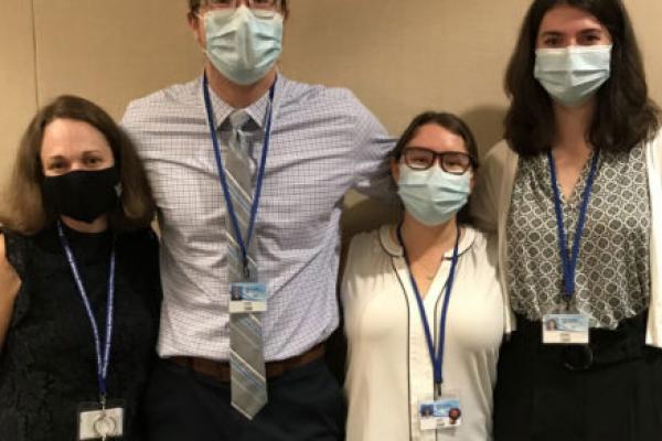 1 man and 3 women are standing in a line with their arms around each other's shoulders. They are all wearing face masks.