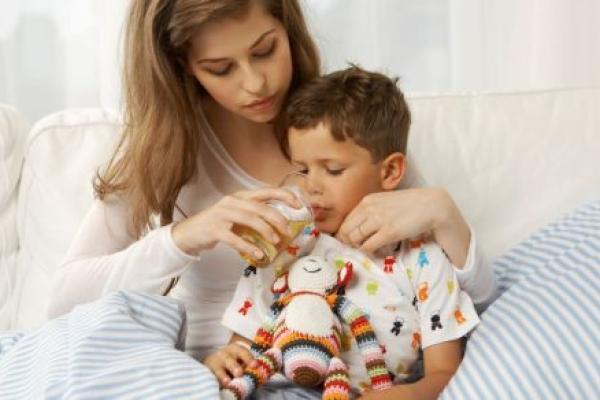 An adult woman holds her son as she helps him drink a glass of juice.