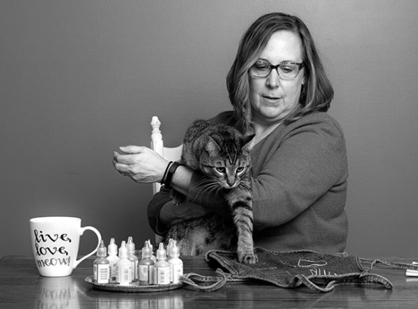 Wendy Johnson, who has shoulder-length hair and wears glasses, sits behind a table and holds her cat, which puts one paw down on a denim apron on the table. Bottles of fabric paint and a mug that says “Live, Love, Meow” are on the table.