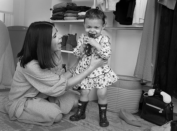 Alyssa Acevedo拥有肩长头发，穿着长袖连衣裙，拥抱她的小孩女儿，他站在她面前。Izzy穿着印花连衣裙和咀嚼太阳镜。鞋子和衣服围绕着衣橱里的架子和地板。